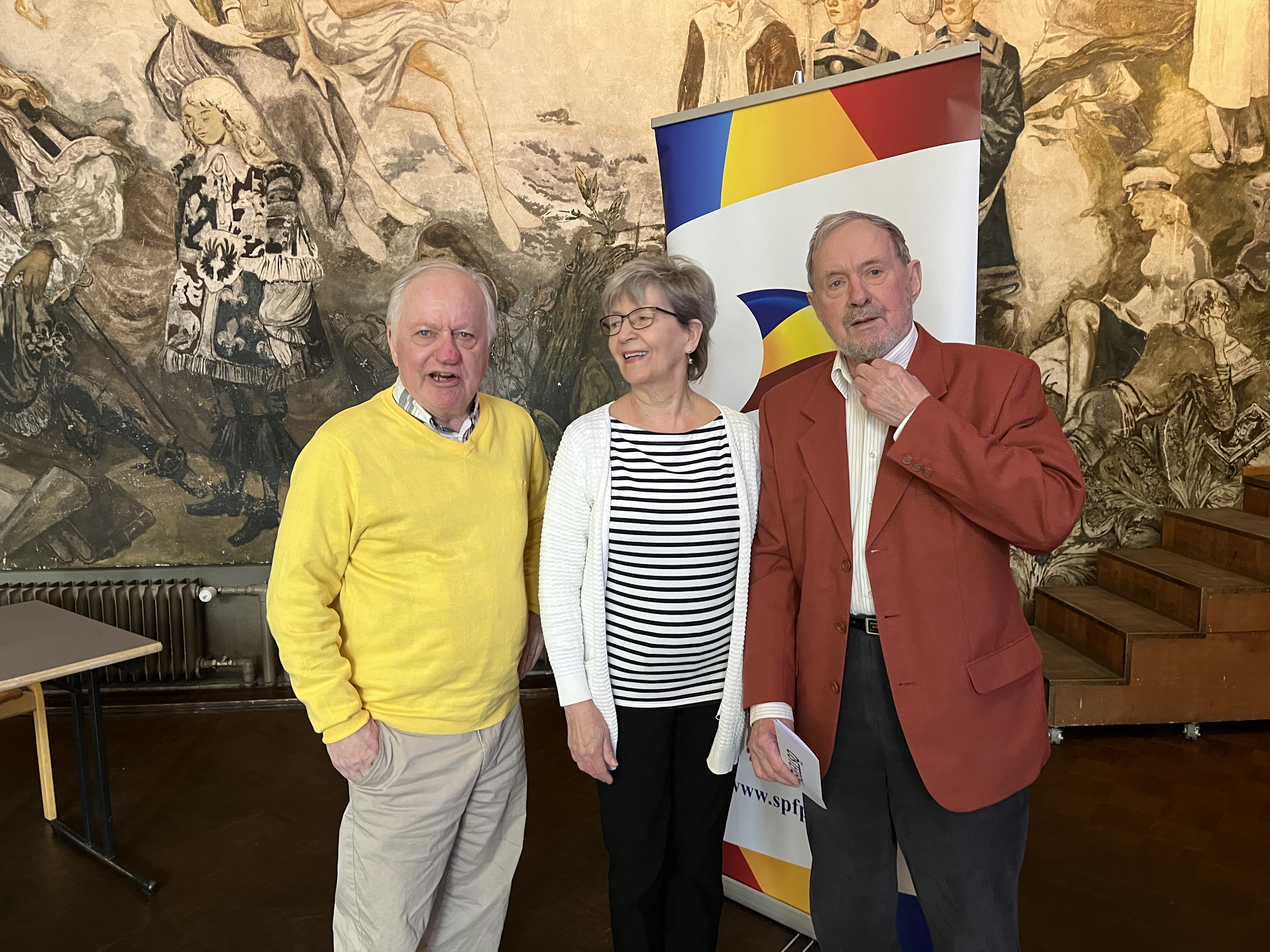 Tre äldre vuxna står inomhus framför en färgglad banderoll och en stor väggmålning. Mannen till vänster bär en gul tröja, kvinnan i mitten bär en randig topp och mannen till höger bär en röd kavaj.