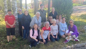 En grupp på fjorton vuxna poserar utomhus i ett skogsparti med träd och solljus i bakgrunden. Några står upp, medan andra sitter på huk eller i gräset. De är klädda i vardagskläder.