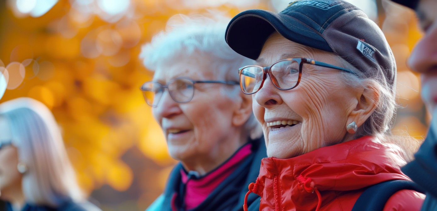 Två äldre kvinnor ler utomhus, båda bär glasögon och kavaj, den ena i röd rock och svart keps. I bakgrunden syns oskarpt höstlövverk i varma nyanser av orange och gult.
