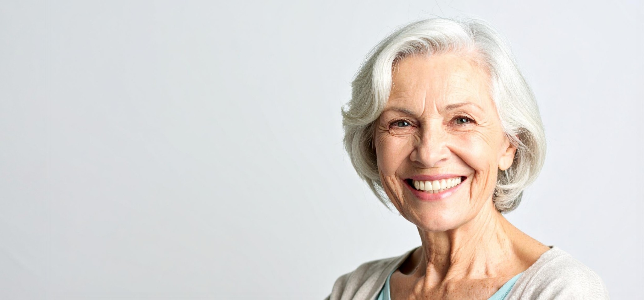 En äldre kvinna med kort grått hår ler mot kameran. Hon är klädd i en ljus topp och kofta och står mot en vanlig ljus bakgrund.