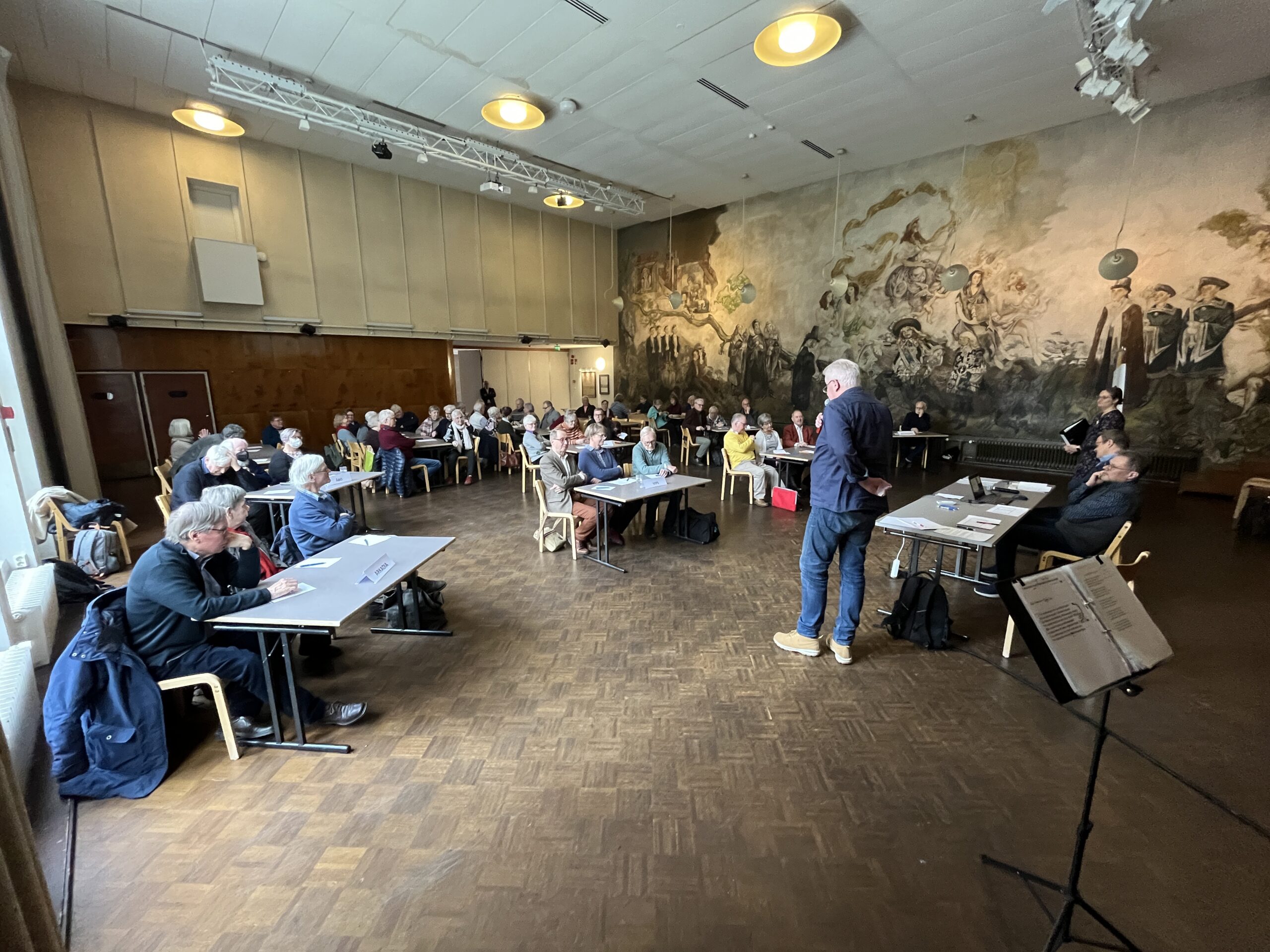 En grupp människor sitter vid bord och stolar i ett stort rum med en väggmålning på ena väggen, medan en man står och talar längst fram. En panel bestående av tre personer sitter till vänster om honom. Stämningen verkar vara uppmärksam.