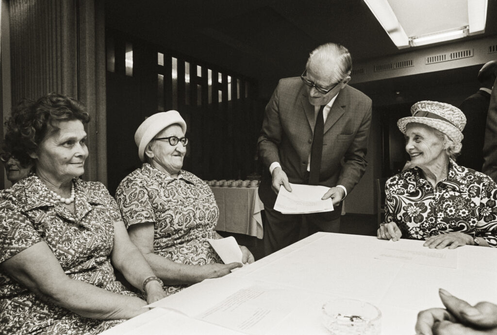 Tre äldre kvinnor sitter vid ett bord, två av dem bär mönstrade klänningar och hattar, medan en man i kostym och glasögon står och lutar sig fram för att ge papper till kvinnan till höger. Miljön ser ut att vara inomhus, möjligen ett möte eller evenemang.