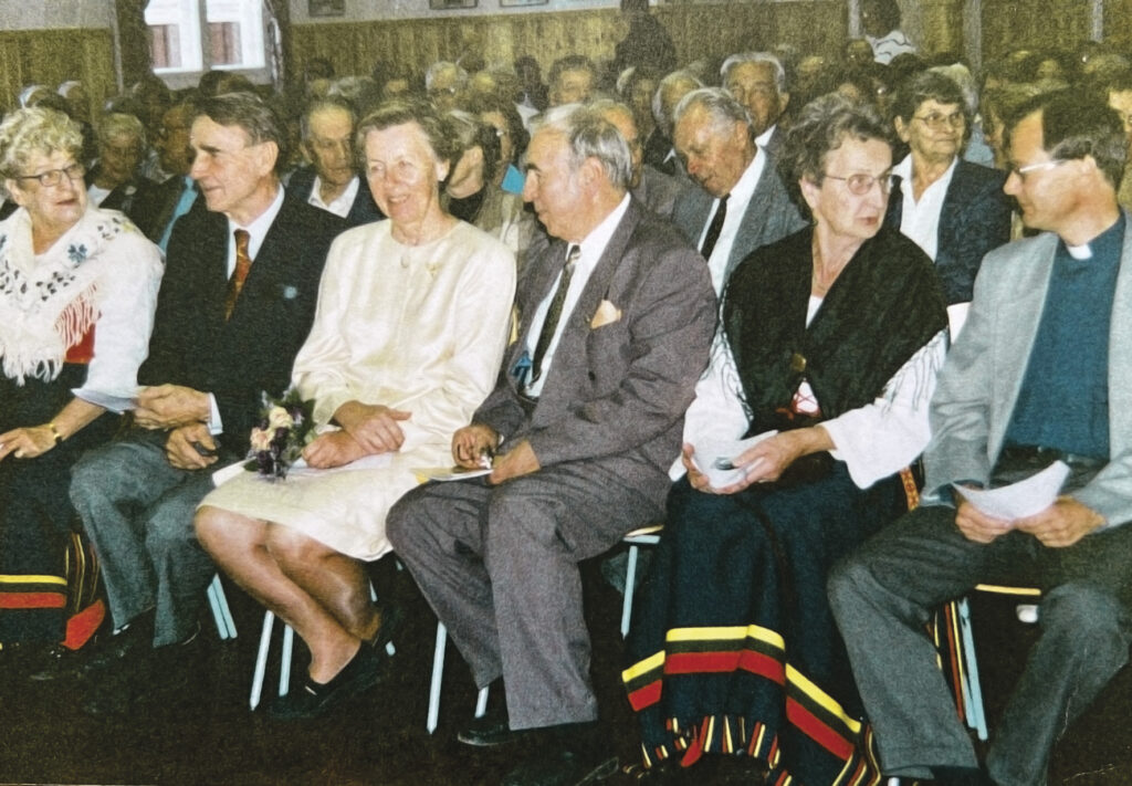 En grupp äldre vuxna sitter på första raden på ett evenemang med många deltagare, klädda i formell klädsel och flera personer bär sjalar med färgglada ränder. Miljön ser ut att vara inomhus med många deltagare.