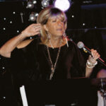 En kvinna med kort blont hår sjunger på scenen i starkt ljus, klädd i en svart dräkt, stora örhängen och ett långt halsband, med ena handen upplyft mot örat.