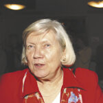 En äldre kvinna med kort vitt hår, klädd i röd jacka, mönstrad scarf och pärlhalsband, tittar lite åt sidan. Hon har en namnbricka och befinner sig inomhus med suddiga människor och lampor i bakgrunden.