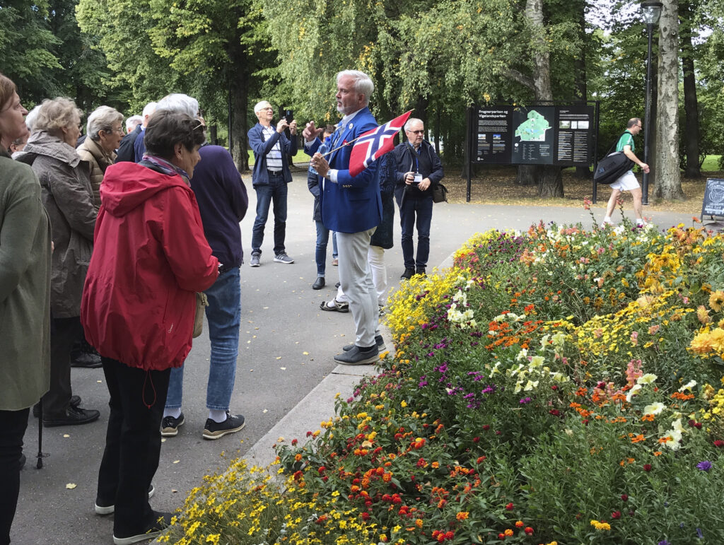 En reseledare som håller i en norsk flagga talar till en grupp människor som står bredvid en färgglad blomsterrabatt i en park med träd och en informationstavla i bakgrunden.