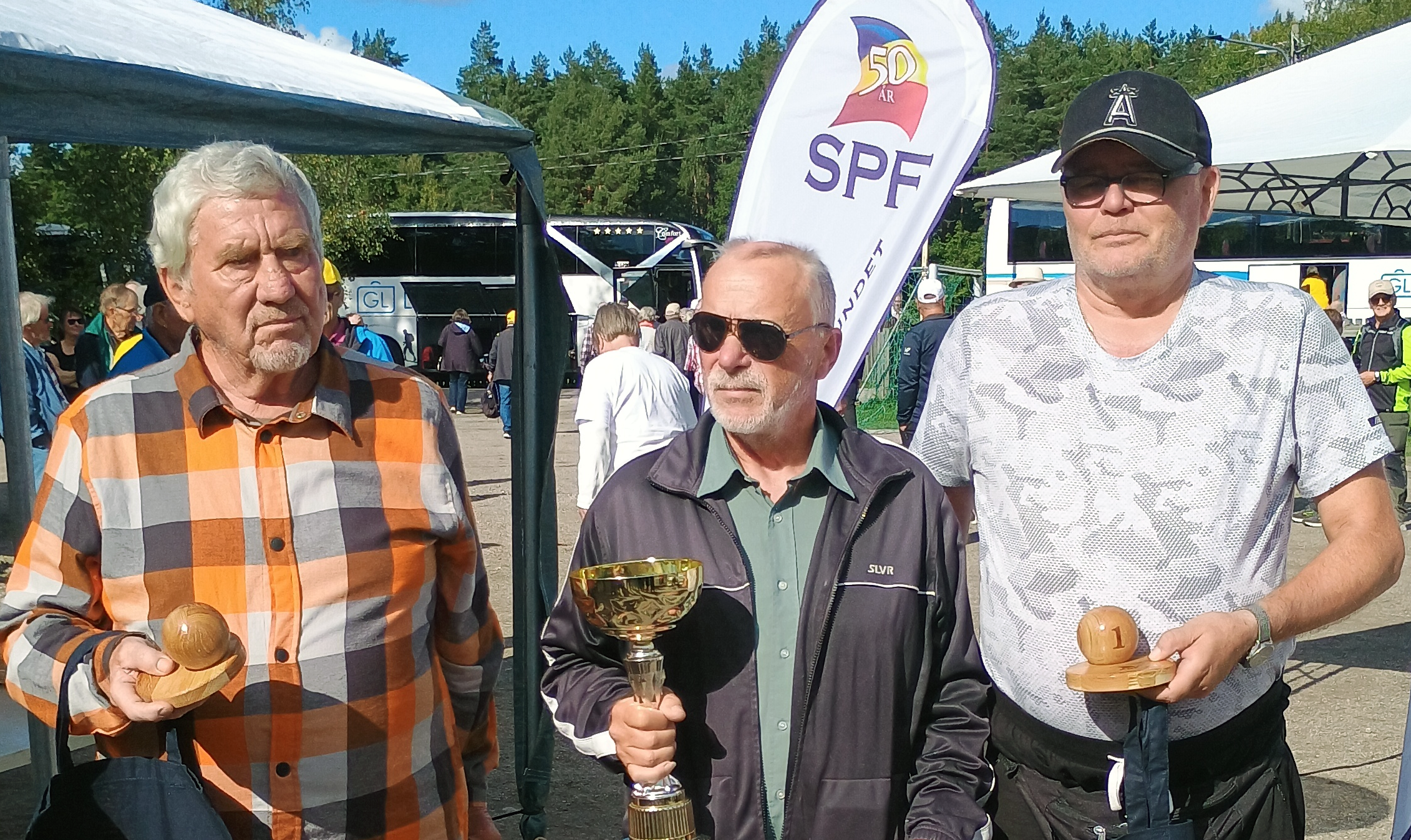 Tre män står utomhus och håller i troféer. Mannen i mitten håller en stor guldpokal, medan de två männen på vardera sidan håller små träpriser. I bakgrunden syns en folkmassa, tält och en SPF-banderoll.