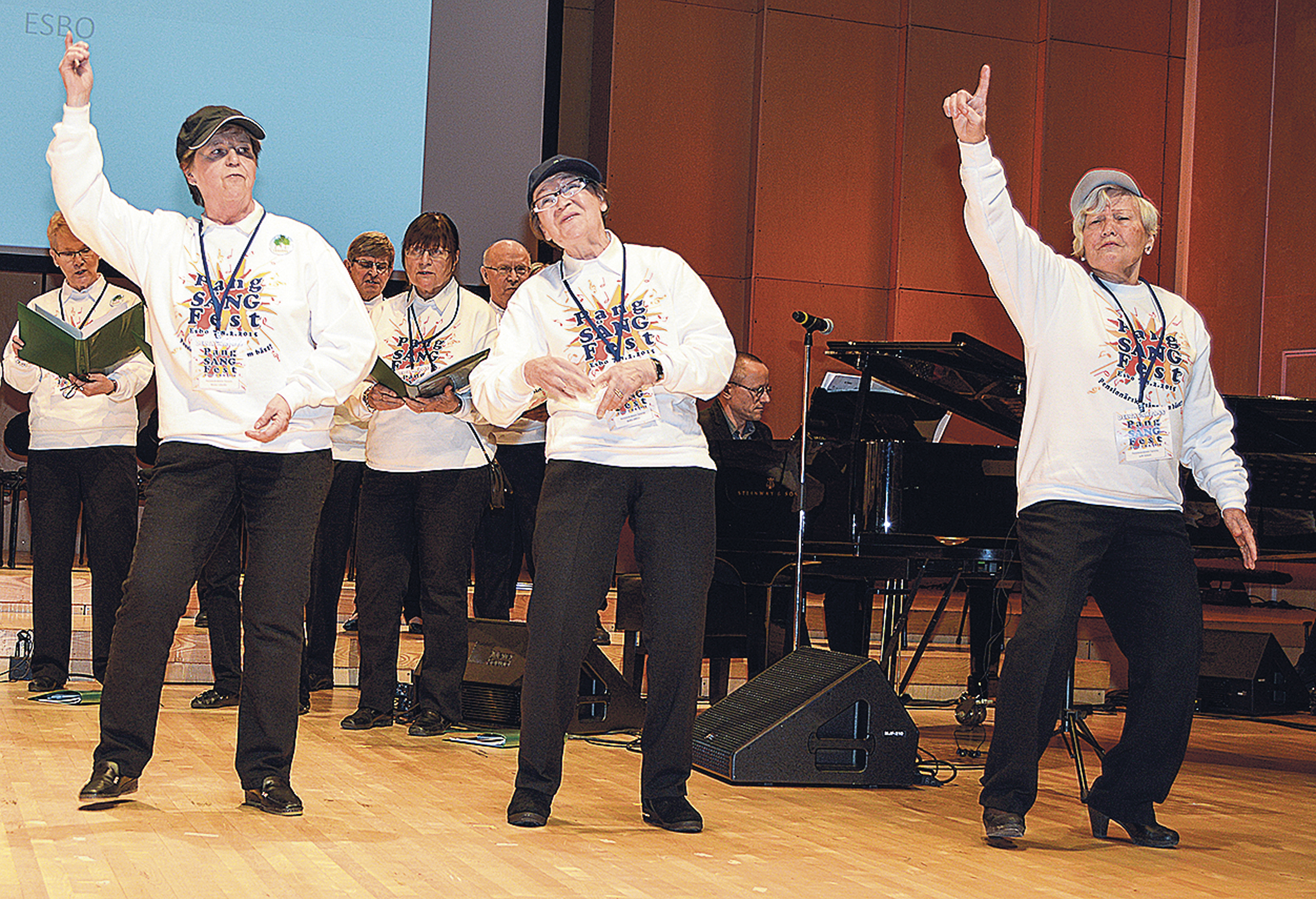 Tre äldre kvinnor i vita sweatshirts och svarta byxor dansar på en scen. Bakom dem står en grupp människor med sångböcker i händerna och en flygel syns. Miljön ser ut att vara en musikalisk föreställning.