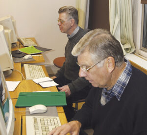 Två äldre män sitter vid skrivbord och använder stationära datorer i ett klassrum eller kontorsmiljö. Papper, anteckningsböcker och mappar ligger på borden och det finns fönster med gardiner i bakgrunden.