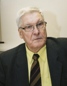 En äldre man med kort vitt hår, glasögon och allvarligt ansiktsuttryck bär en mörk kostymkavaj, en ljusgul skjorta och en randig slips. Han sitter framför en ljus bakgrund.