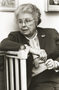 En äldre kvinna med kort, lockigt hår och glasögon sitter bredvid ett räcke, klädd i kostymkavaj, blus med rosett och brosch. Hon vilar en arm på räcket och tittar åt sidan i en hemmiljö.