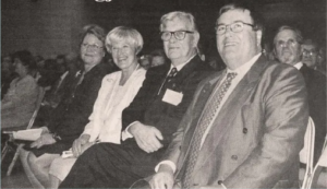 Fyra vuxna i formell klädsel sitter på rad på ett inomhusevenemang. De är vända framåt och ler. Texten ovanför dem på svenska lyder: Ni har gjort det bra för oss (You have done well for us).