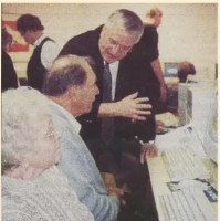 En äldre man och kvinna sitter vid en dator medan en annan man, som står bredvid dem, gestikulerar mot bildskärmen och talar till dem. Flera andra personer syns i bakgrunden vid datorer.