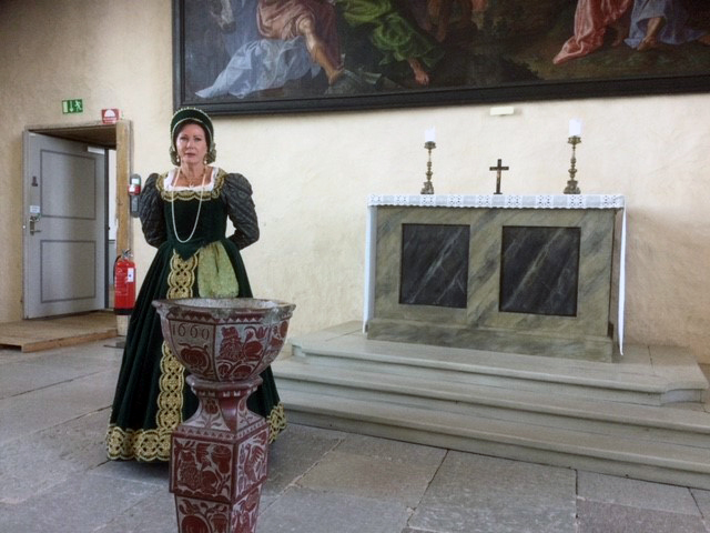 En kvinna i grön klänning i renässansstil står vid en dopfunt i en kyrka. Bakom henne finns ett altare med två ljus, ett kors och en stor religiös målning på väggen.