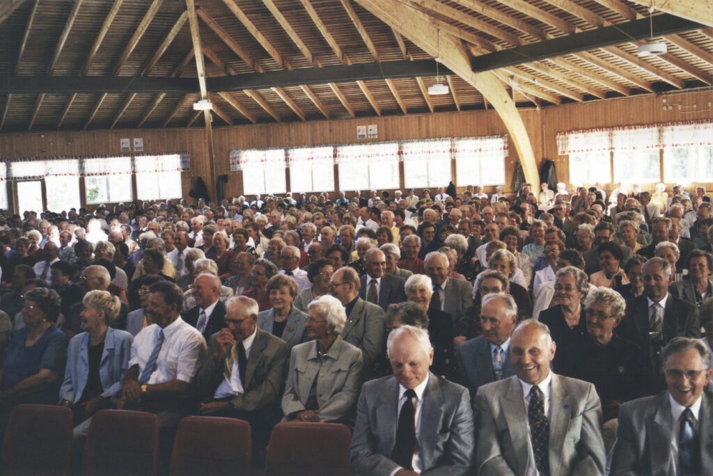 En stor grupp människor, mestadels äldre vuxna, sitter tätt tillsammans i rader i en trähall med synliga bjälkar och stora fönster, vända mot kameran och ler.