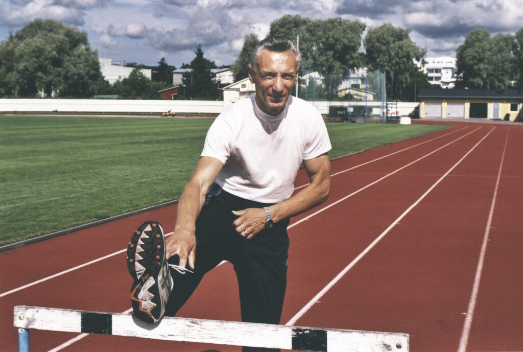 En äldre man i vit t-shirt och mörka byxor sträcker ut benet på en häck på en löparbana utomhus, med grönt gräs, byggnader och träd i bakgrunden under en delvis molnig himmel.