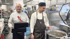 En äldre man med två svarta matbehållare i handen står bredvid en kock som rör om i en stor gryta i ett storkök med industriell matlagningsutrustning.