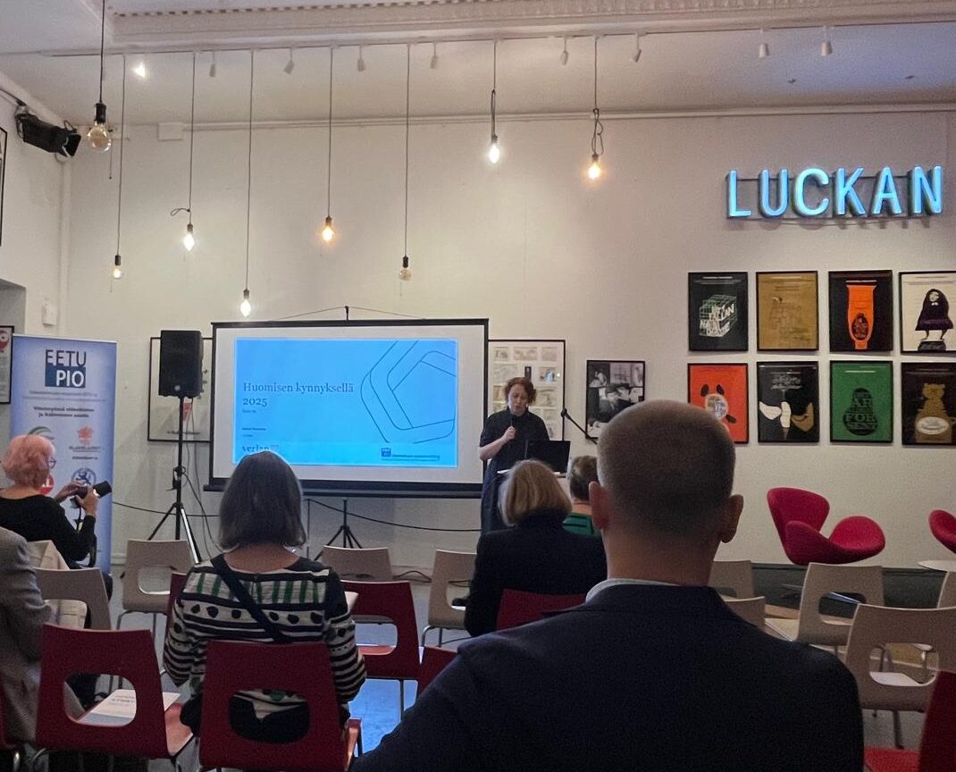 En presentatör står nära en projektionsskärm som visar en bild i ett litet rum med en publik som sitter på röda stolar. På väggen bakom syns inramade affischer och en LUCKAN-skylt i neon. Hängande lampor lyser upp scenen.