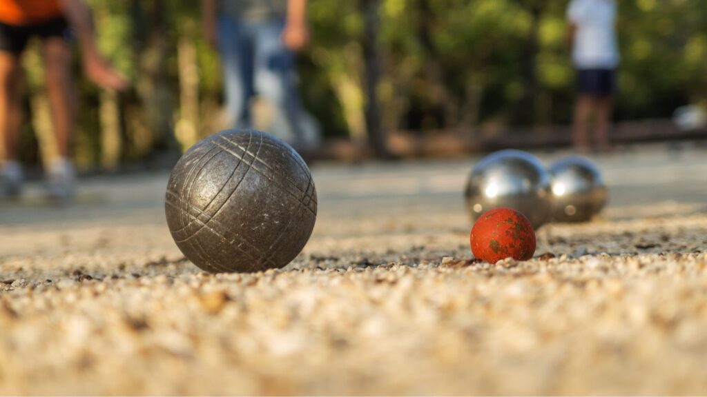 Närbild av ett metalliskt bouleklot och ett litet rött målklot på en grusyta, med andra bouleklot och suddiga människor i bakgrunden.