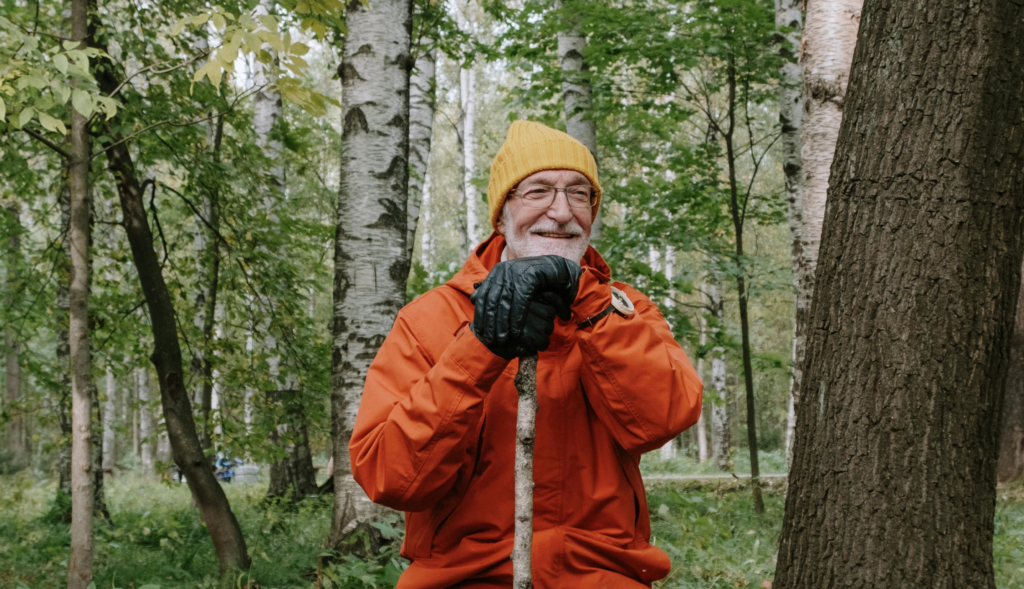 En äldre man med vitt skägg och glasögon sitter i en skog, klädd i en gul stickad mössa, orange jacka och svarta handskar, håller i en käpp och ler. Björkar och grönt lövverk omger honom.