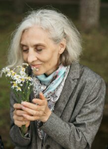 En äldre kvinna med grått hår sitter på en gunga utomhus och håller i och luktar på en liten bukett med vita vildblommor. Hon är klädd i en grå kappa och en mönstrad scarf. Träd och gräs syns i bakgrunden.