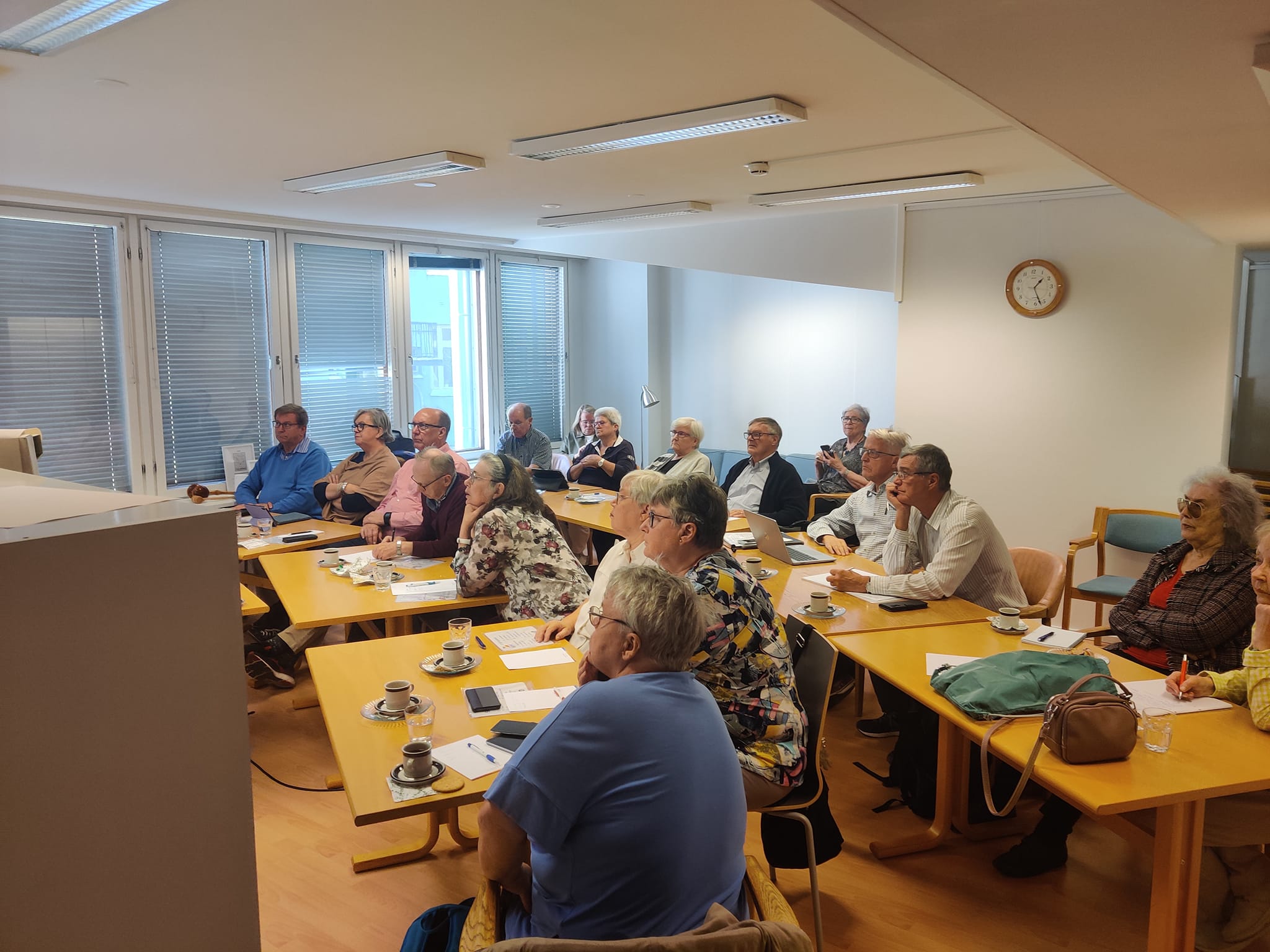 En grupp vuxna sitter runt bord i ett mötesrum, vända mot en skärm eller högtalare utanför bild. Anteckningsböcker, kaffekoppar och papper ligger på borden. Rummet har stora fönster och en klocka på väggen.