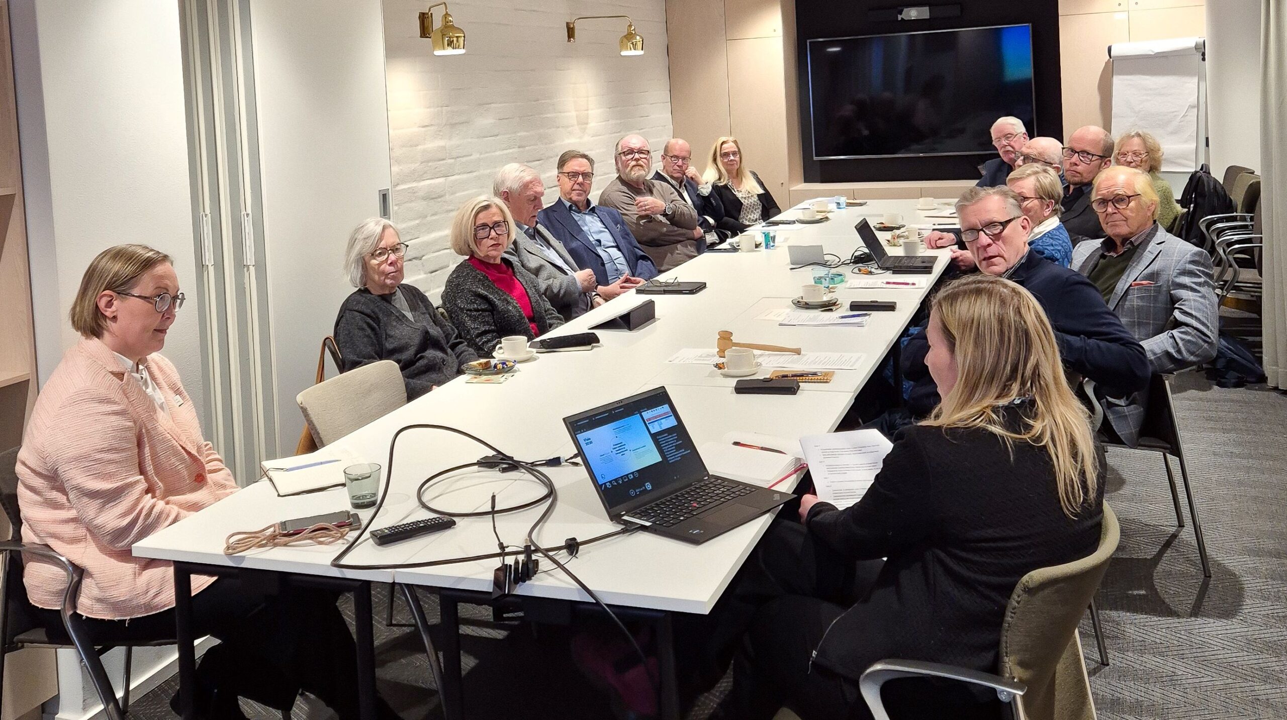 En grupp människor, mestadels äldre vuxna, sitter runt ett konferensbord i ett mötesrum. Papper, bärbara datorer och kablar ligger på bordet.