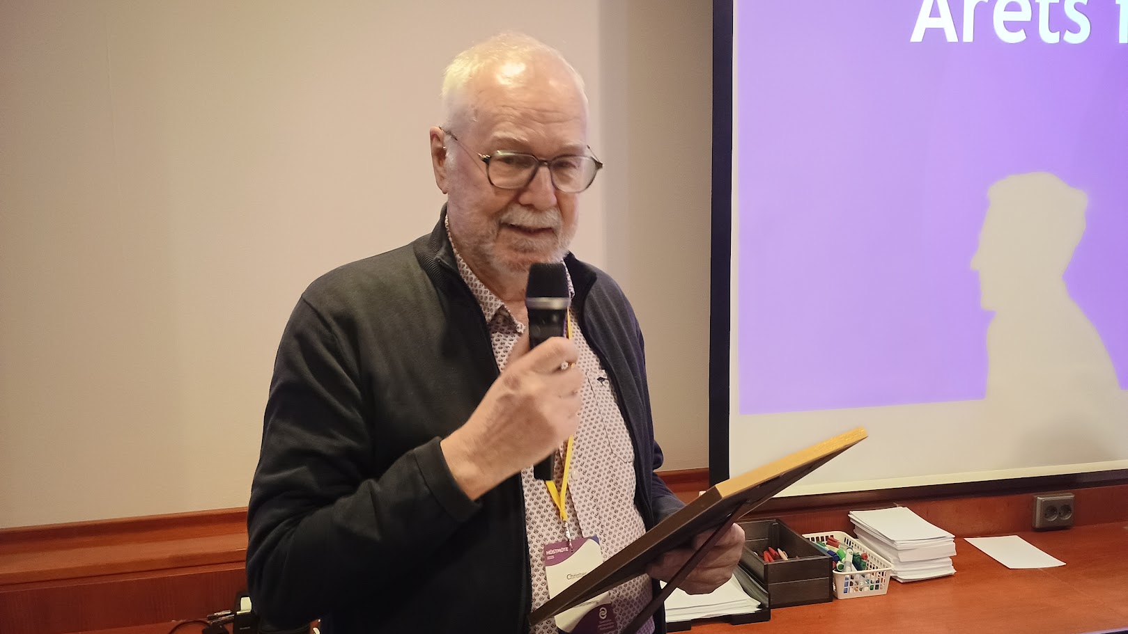 En äldre man med mikrofon och plakett står framför en skärm som visar text. Han bär glasögon, ett nyckelband och en mörk jacka. På ett bord bredvid honom ligger papper och små lådor.