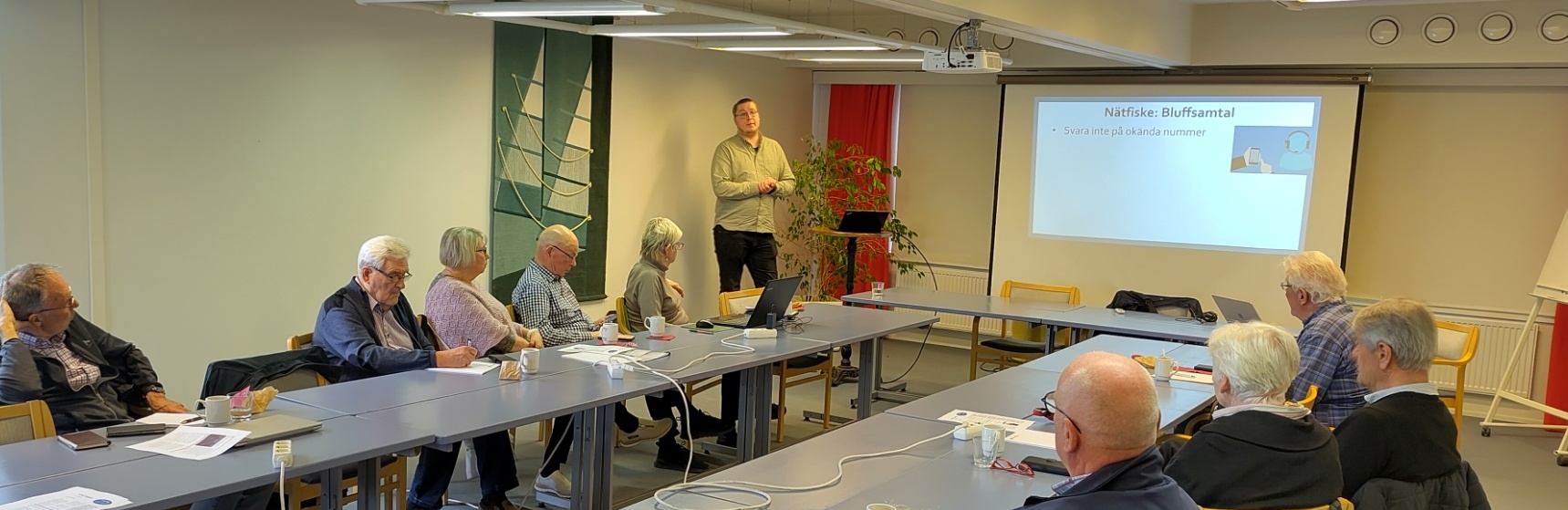 En grupp äldre vuxna sitter runt bord i ett mötesrum och lyssnar på en man som står vid en skärm som visar en presentationsbild med titeln Nätkafé, Blufsamtal. På borden finns papper, dryck och en bärbar dator.