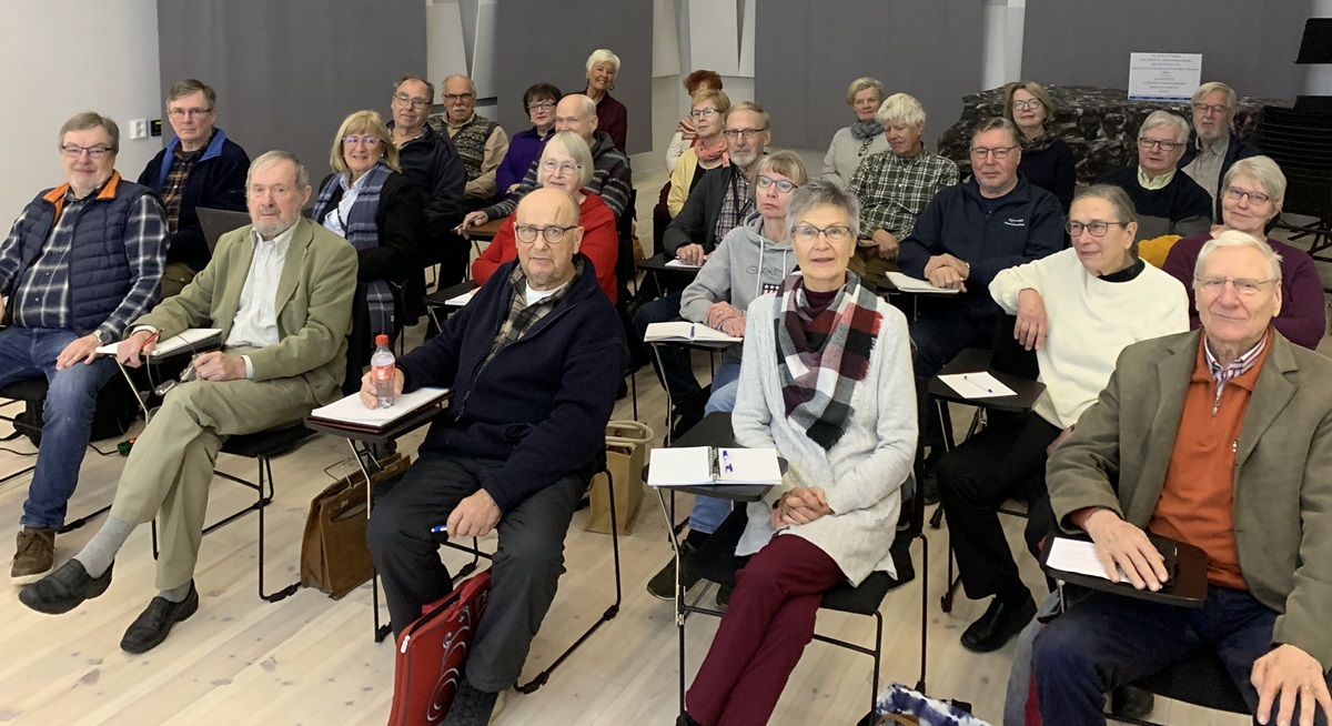 En grupp vuxna och äldre sitter framåtvända i ett klassrum med anteckningsblock och pennor i knäna. Rummet har ljust golv, svarta stolar och gråa väggar. De flesta deltagarna bär glasögon och vardagskläder.
