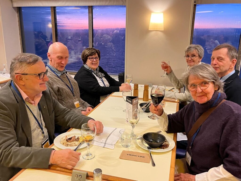 Sex äldre vuxna sitter runt ett bord med mat och dryck i en matsal och ler mot kameran. Ett fönster bakom dem visar en solnedgång över havet. På bordet finns tallrikar, glas och en skylt med texten Viking Line.