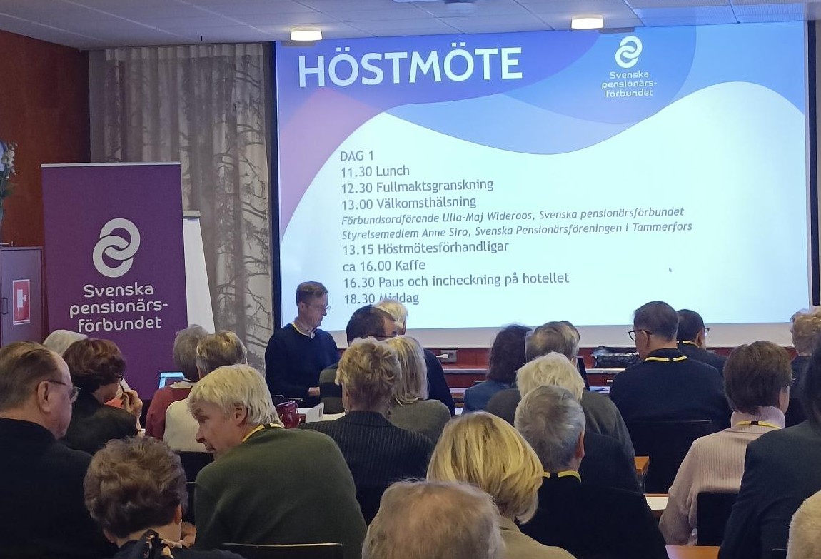 En grupp människor sitter framför en skärm på Svenska pensionärsförbundets höstmöte. Dagordningen visas på skärmen och två talare sitter vid ett bord framför publiken.