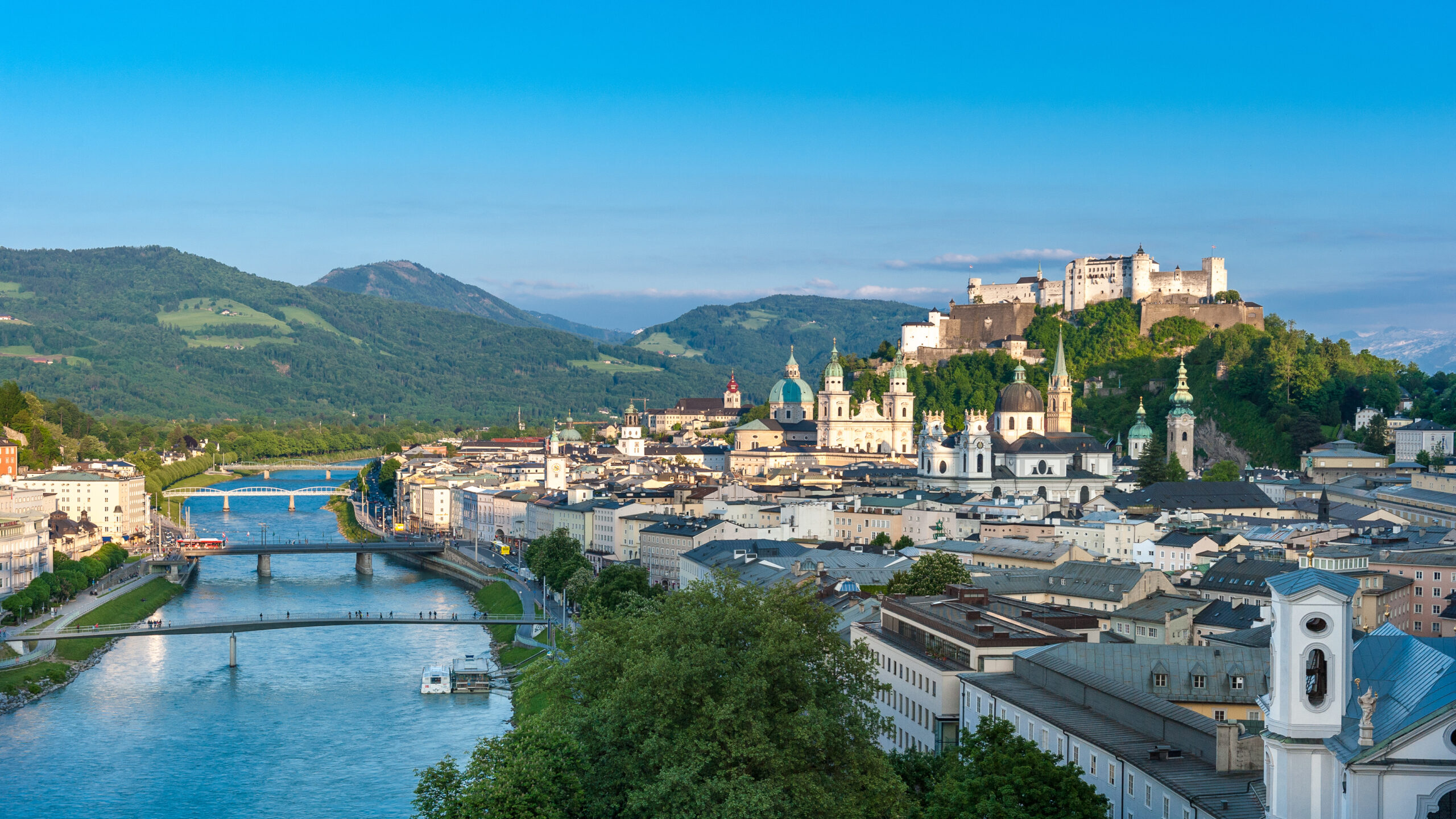 Panoramavy över Salzburg i Österrike med floden Salzach, flera broar, historiska byggnader, gröna kullar och fästningen Hohensalzburg på en kulle under en klarblå himmel.