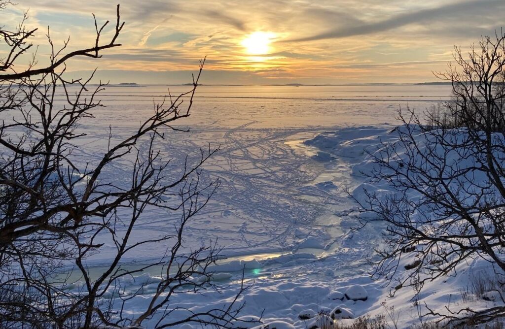 Snötäckt, fruset landskap med delvis molnig himmel i solnedgången. Nakna trädgrenar ramar in förgrunden, medan spår och sprickor syns på den isiga ytan som sträcker sig till horisonten.