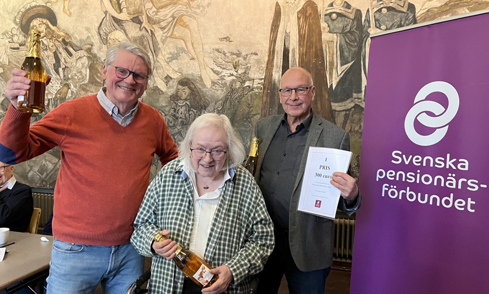 Tre äldre vuxna står inomhus framför en väggmålning och en lila banderoll med texten Svenska pensionärsförbundet. Två håller i flaskor och en håller i ett diplom, alla ler.
