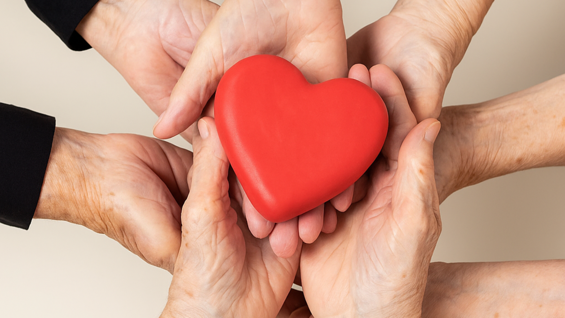 Flera par äldre händer håller ihop ett rött hjärtformat föremål, vilket symboliserar omsorg, stöd och enighet. Bakgrunden är neutral och drar fokus till händerna och hjärtat.