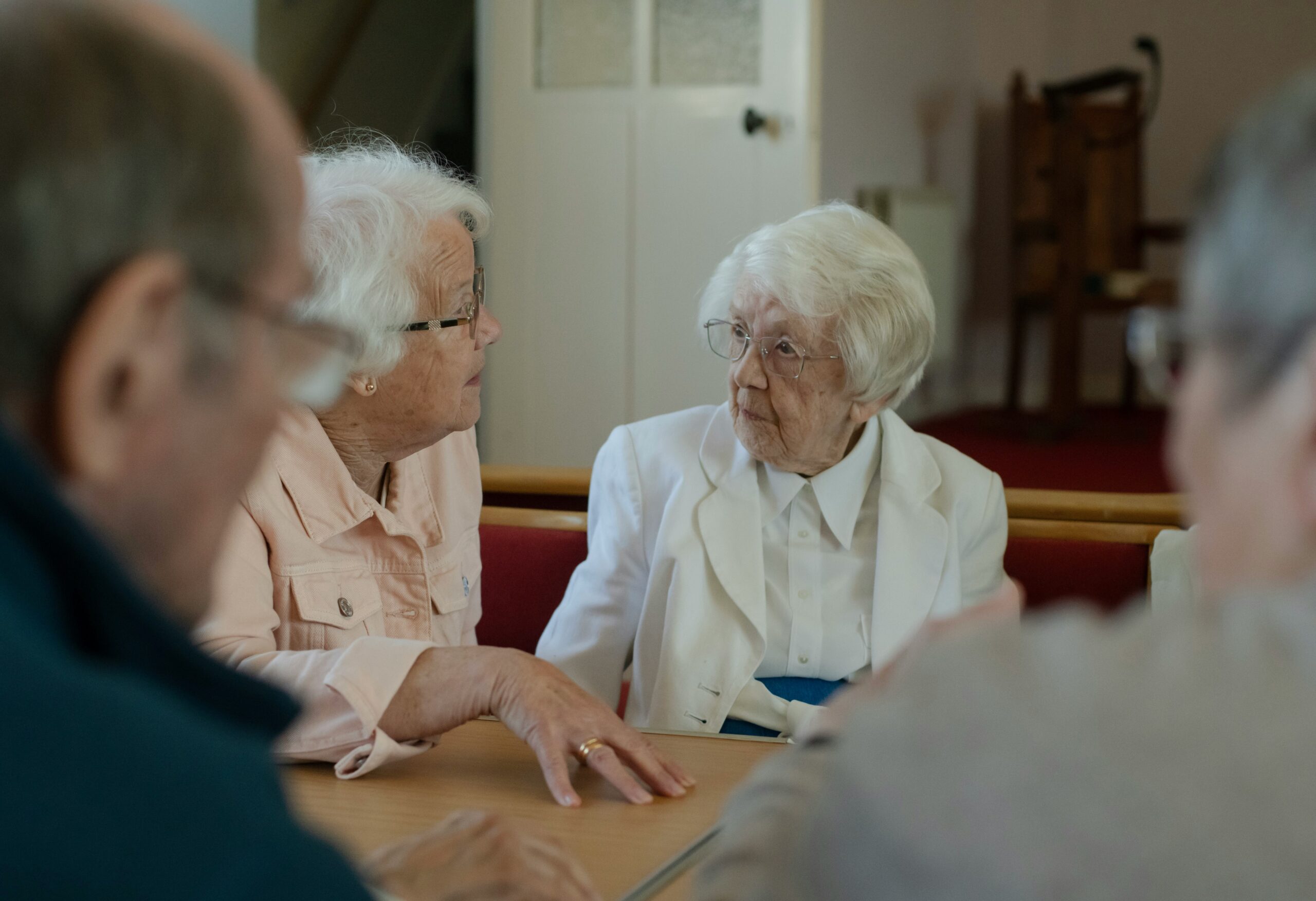 Två äldre kvinnor sitter vid ett bord i ett mötesrum och samtalar, med två andra personer suddiga i förgrunden.