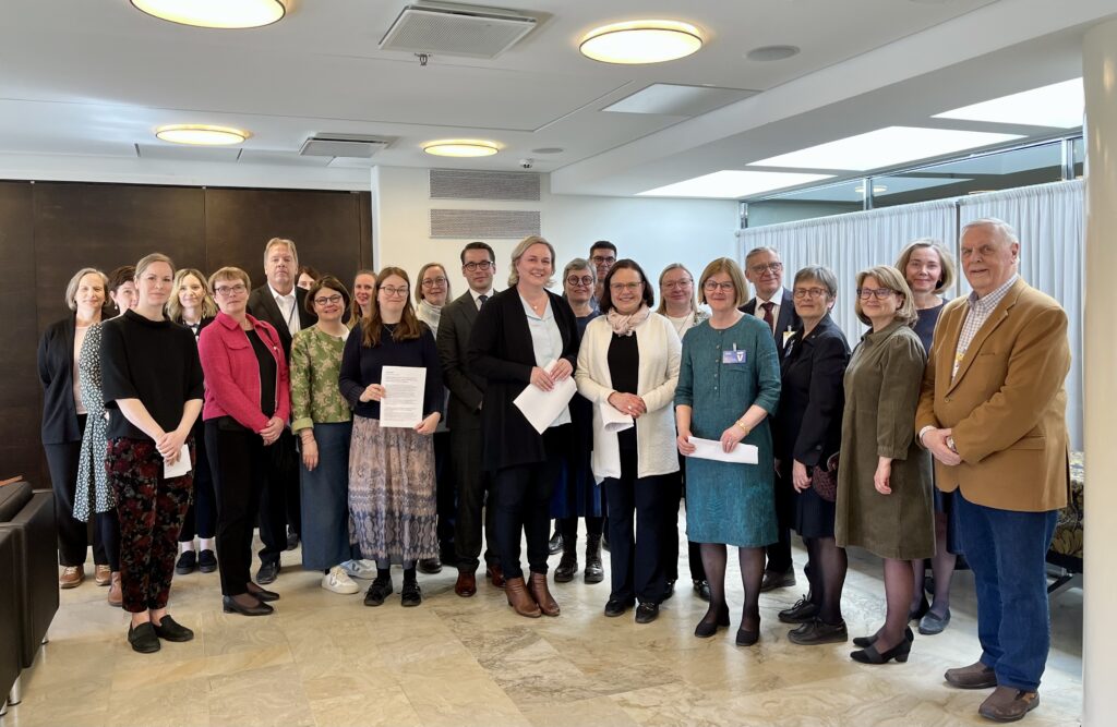 En grupp på cirka 22 vuxna står inomhus och poserar och ler för ett gruppfoto. Flera personer längst fram håller i papper. Miljön ser ut att vara ett konferensrum eller kontor med stark belysning.