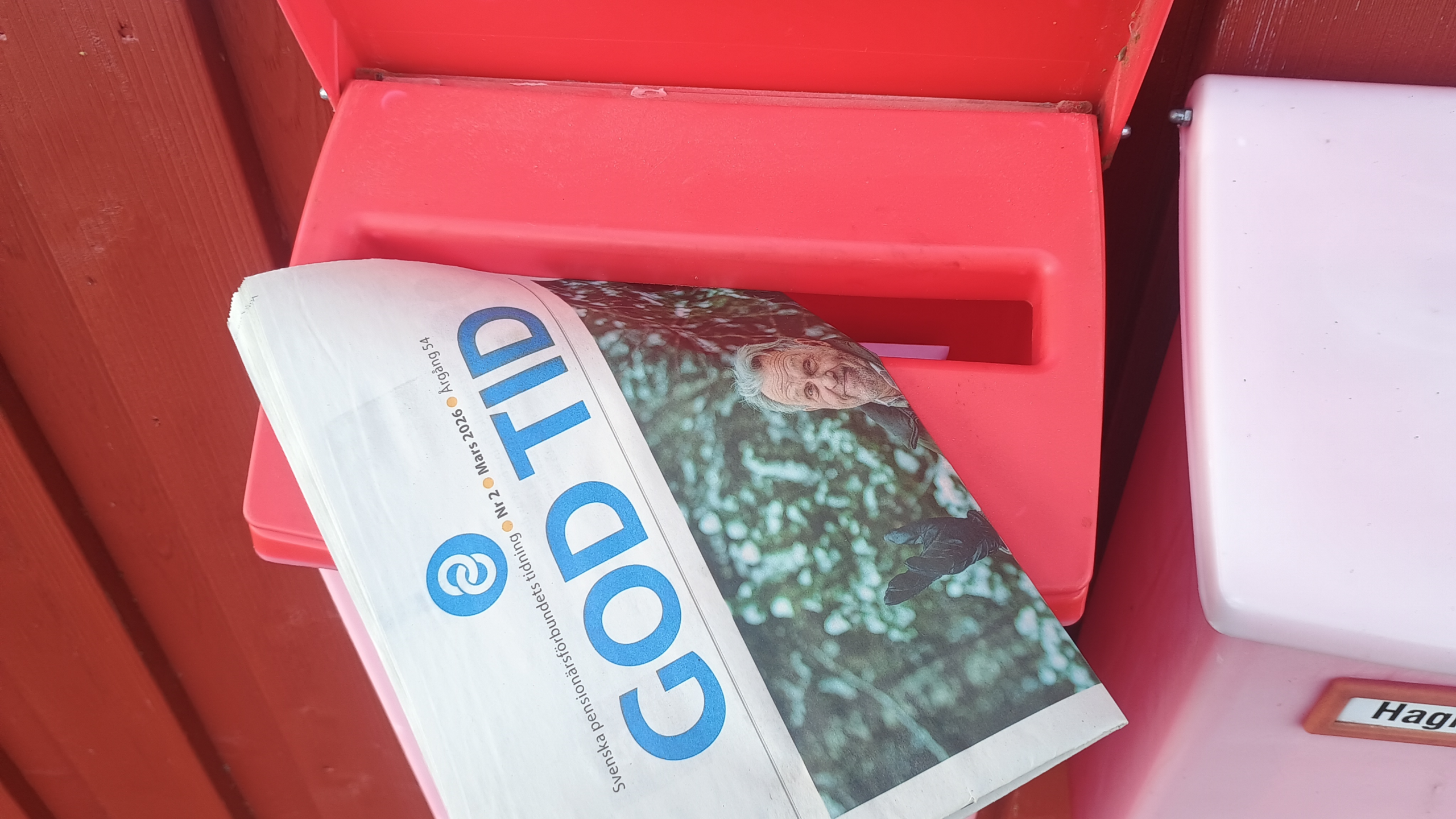 En hoprullad tidning med rubriken GOD TID är placerad i facket på en röd brevlåda som är fäst vid en trävägg. En del av en andra behållare märkt Hag... syns till höger.