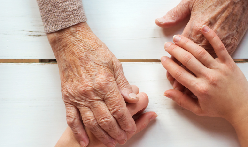 Närbild av två äldre händer som försiktigt håller en yngre persons hand på en vit träyta, vilket framhäver kontrasten i hudstruktur och ålder.