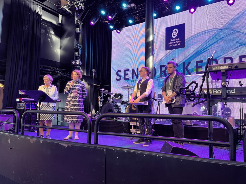 Fyra personer står på en scen med mikrofoner och musikutrustning bakom sig. En stor skärm i bakgrunden visar orden Senatorekade med en blå abstrakt design och logotyper. Scenen är upplyst av scenljus.