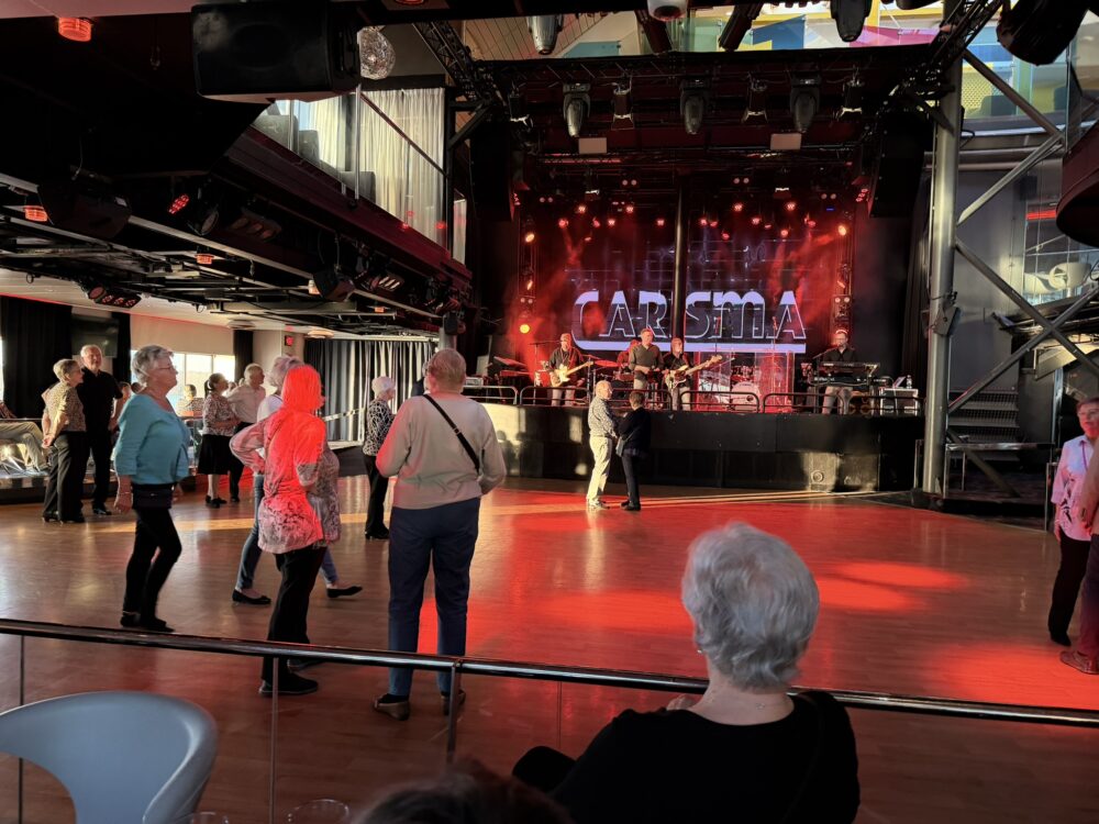 Människor dansar på ett trägolv framför en scen med röd belysning, där ett band uppträder under en stor skylt med texten CARISMA. Flera personer står och tittar på, och stolar syns i förgrunden.