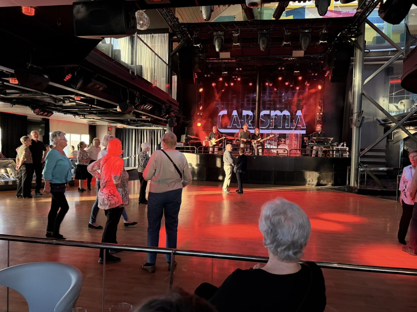 Människor dansar på ett trägolv framför en scen med röd belysning, där ett band uppträder under en stor skylt med texten CARISMA. Flera personer står och tittar på, och stolar syns i förgrunden.