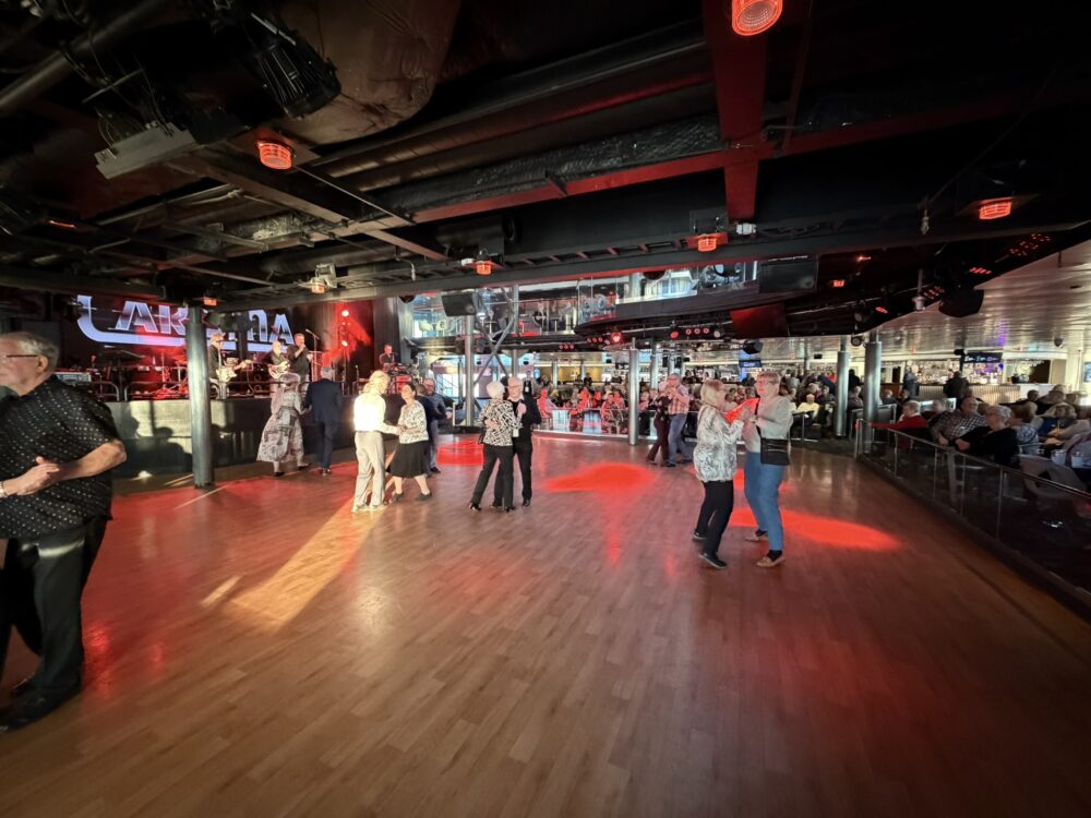 Människor dansar på ett trägolv i en stor inomhuslokal med röd belysning. Ett band spelar på scenen till vänster, medan andra människor sitter vid bord i bakgrunden och tittar på. Några fönster släpper in naturligt ljus.