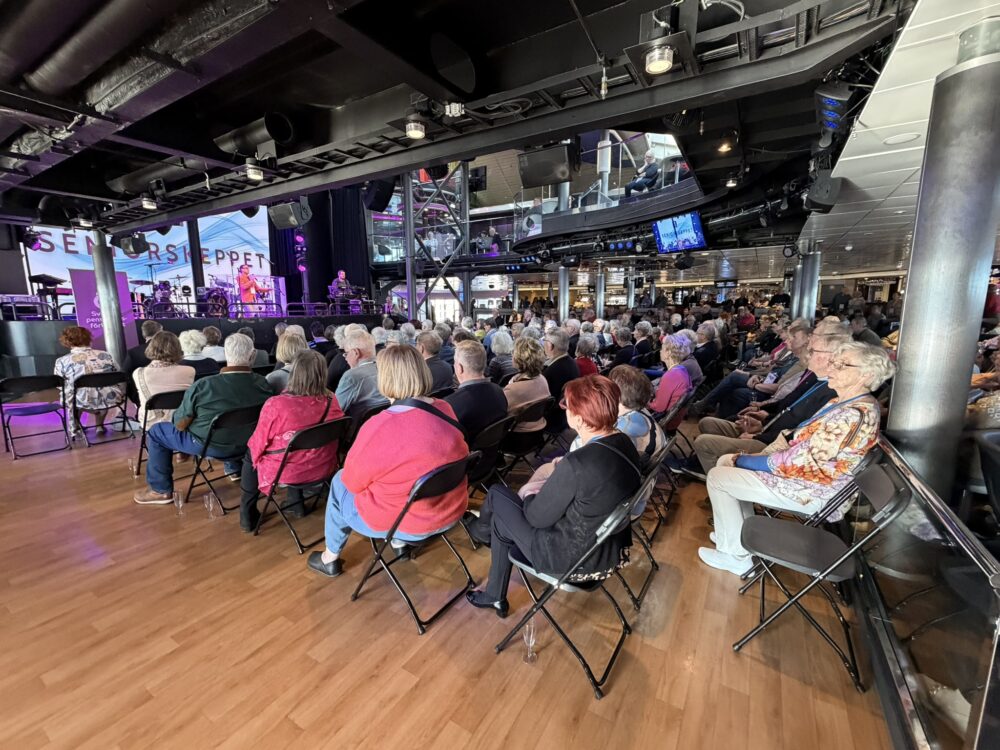 En stor grupp människor sitter på fällstolar i en inomhuslokal och tittar på ett liveband som uppträder på en scen. Rummet har trägolv, metallbalkar och olika skärmar som visar evenemanget.
