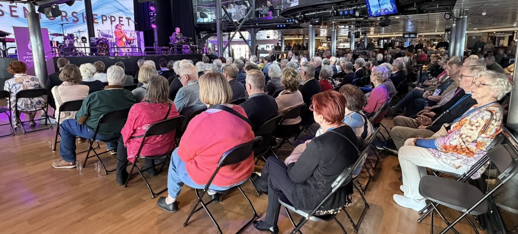 En stor publik bestående av främst äldre sitter på fällstolar inomhus och ser ett liveband uppträda på en scen med Seniors Steppet skrivet i bakgrunden. Lokalen är väl upplyst och rymlig.