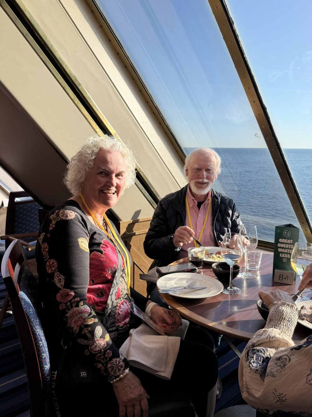Två äldre vuxna sitter vid ett bord vid ett stort fönster med utsikt över havet och äter en måltid med dryck. Solljuset skiner in och båda bär gula nyckelband. Tallrikar, glas och en meny står på bordet.