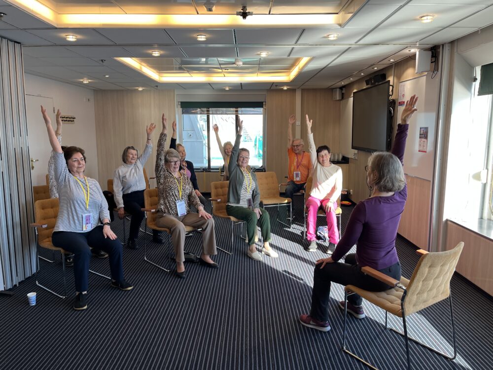 En grupp äldre personer, som sitter på stolar i en halvcirkel i ett konferensrum, lyfter en arm var medan de vänder sig mot en instruktör längst fram i rummet. Rummet har stark belysning och stora fönster.