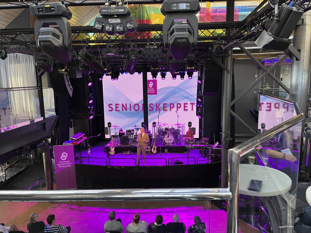 En scen med musikinstrument och en storbildsskärm som visar Senior Skeppet är uppställd i en modern lokal. Flera personer sitter i publiken och en person står på scenen. Belysningsutrustning hänger ovanför.