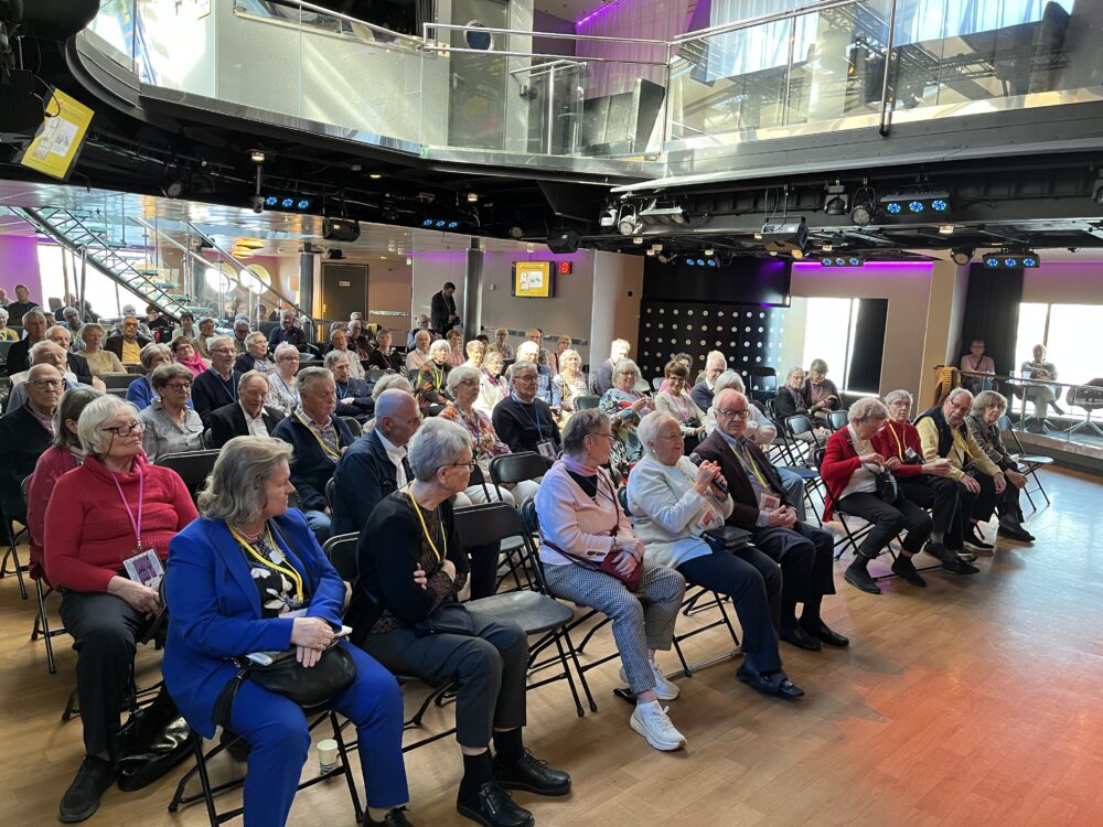 En grupp äldre personer sitter på fällstolar i ett rymligt, modernt rum med trägolv och stark belysning, vända framåt som om de skulle delta i en presentation eller ett evenemang. Några personer pratar eller håller i mikrofoner.